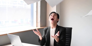 一位亚洲华裔年轻白领坐在办公室的椅子上，把文件扔向空中，心情沮丧，情绪紧张