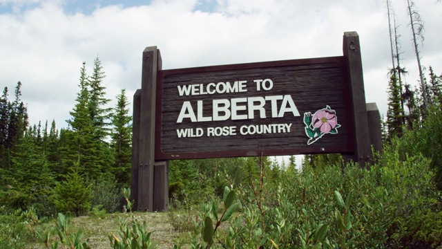 在一个多云的日子里，一辆汽车沿着美国/加拿大边境进入阿尔伯塔省，路边有“欢迎来到阿尔伯塔，野生玫瑰之乡”的标志