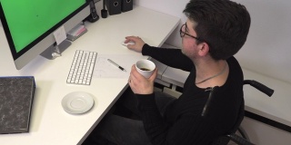 坐着轮椅的办公室职员，深色头发的女人在电脑旁边的工作场所喝咖啡，慢镜头