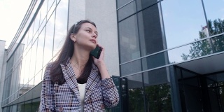 一个穿着休闲服的年轻女孩站在商业中心的背景上，手里拿着一部现代手机。商务女性在她的智能手机上输入信息。办公室附近的电话沟通