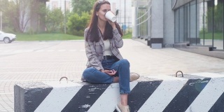 女孩坐在混凝土分隔块上，用纸杯喝热咖啡或茶