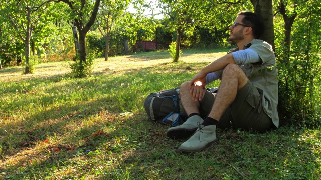 一个人在树下享受着温暖阳光的夏日，在他的徒步旅行中休息