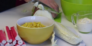 一个男人在做北京大白菜沙拉。在菜板上用刀切卷心菜。桌子旁边是其他的食材。玉米、蟹肉条和蛋黄酱。特写镜头