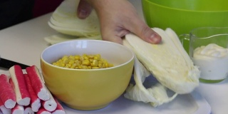 一个男人在做北京大白菜沙拉。在菜板上用刀切卷心菜。桌子旁边是其他的食材。玉米、蟹肉条和蛋黄酱。特写镜头。