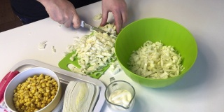 一个男人在做北京大白菜沙拉。用菜板上的刀子把卷心菜切成片，然后扔进一个容器里。桌子旁边是其他的食材。玉米、蟹肉条和蛋黄酱