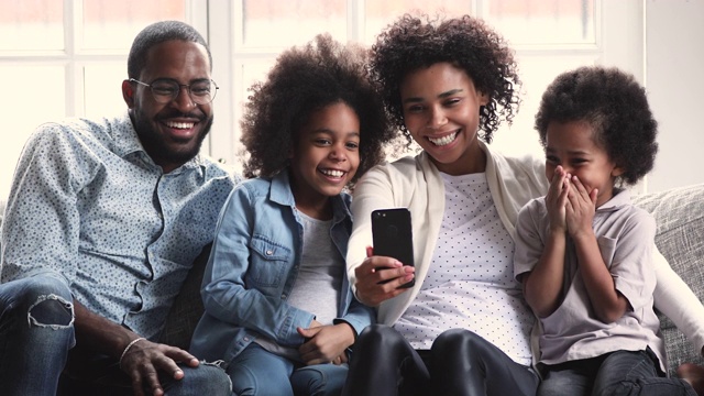 快乐的非洲父母和孩子们在沙发上用手机大笑