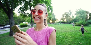 微笑的女人与滑稽的脸和粉红色的眼镜站在日落城市绿色公园的背景和使用手机