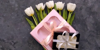 白色郁金香花与黑色和粉红色的礼盒在黑暗的背景平躺。4K视频俯视图。三月八日妇女节快乐的概念。花束，丝带蝴蝶结礼物，女性博客
