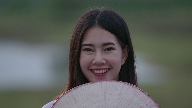 一位美丽的年轻女子穿着一件白色的连衣裙，拿着一顶竹编织的帽子。站着对着镜头笑，心情很好