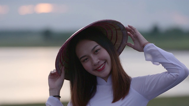 一位美丽的年轻女子穿着一件白色的连衣裙，拿着一顶竹编织的帽子。站着对着镜头笑，心情很好