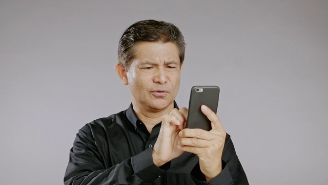 资深男士浏览手机，心烦意乱，孤立在灰色背景中。穿着黑衬衫的亚洲人。