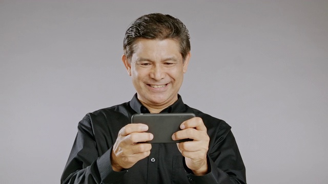 老人在玩他的手机游戏，微笑，胜利，孤立在灰色的背景。穿着黑衬衫的亚洲人
