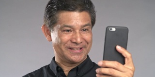 高级男子用手机视频通话，笑着，孤立在灰色背景中。穿着黑衬衫的亚洲人。