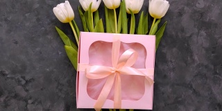 白色郁金香花和粉红色礼盒在深灰色的背景平lay。4K视频俯视图。三月八日妇女节快乐的概念。春天花束，丝带蝴蝶结送礼