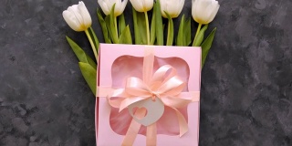 白色郁金香花与粉色礼盒和心卡标签在黑暗的背景平lay。4K视频8三月妇女节快乐的概念。春天鲜花花束
