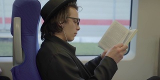 年轻帅气的男子戴着黑帽子和眼镜坐在公共交通工具上看书，千禧潮的学生喜欢在地铁里看书