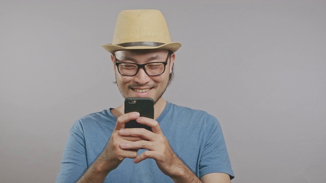 年轻的潮人看手机屏幕孤立在灰色背景。一个戴着帽子和眼镜的亚洲人站在镜头前。中景镜头。