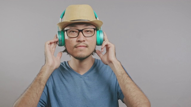 年轻的潮人在灰色背景下用耳机听音乐。一个戴着帽子和眼镜的亚洲人站在镜头前。中景镜头。