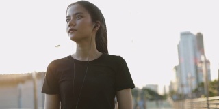 美丽的亚洲女子运动员跑步休息后，在城市的城市日落有氧运动。慢跑的生活方式健康。女孩跑步戴耳机听音乐。