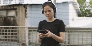 美丽的亚洲女运动员跑步戴着耳机，用智能手机听着音乐在城市里慢跑。慢跑的生活方式健康。