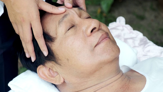 老人在户外做头部按摩。亚洲男人躺在床上。脸部特写。高级生活方式。