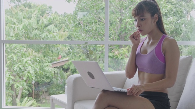 Fit woman使用笔记本电脑，打字，大惊喜，胜利。美丽的亚洲女人在体育。坐在客厅的沙发上。
