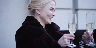 正面美丽的白人金发女人和女性朋友在户外喝香槟的肖像。微笑的女人穿着黑色优雅的外套庆祝。财富、休闲的生活方式,。缓慢的运动。
