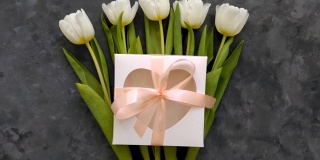 白色郁金香花，心形礼盒与粉色丝带蝴蝶结在深灰色背景平躺4K视频俯视图。母亲节