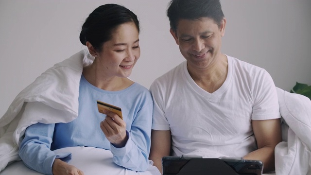亚洲老年夫妇在床上用平板电脑网购。持有金卡信用卡。