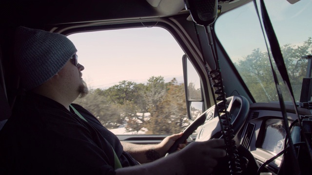 在一个阳光明媚的冬日里，一位三十多岁留着胡子的白人男性卡车司机驾驶着他的半卡车行驶在犹他州高沙漠的一条路上