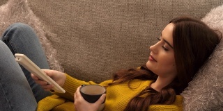 看电视换频道。年轻女子躺在沙发上，手里拿着一个咖啡杯。