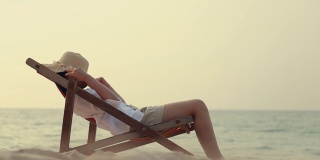 年轻的亚洲女性感觉放松躺在沙滩上的躺椅上有软呢帽接近她的脸。夏天度假的概念。