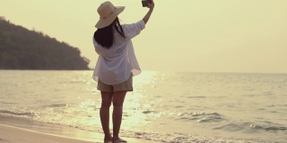 亚洲美丽的年轻女子使用智能手机自拍，而站在海滩日落。生活方式旅游度假夏季概念。