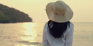 迷人的亚洲女人戴着软呢帽站在海滩上看海景美丽的日落。生活方式旅游度假夏季概念。