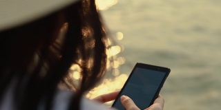 近距离的亚洲女人手打字智能手机上的信息，而站在海滩上的日落。生活方式旅游度假夏季概念。