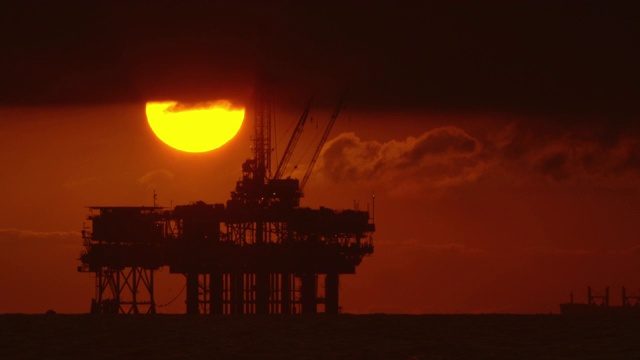太阳从风暴云下与一个剪影的近海石油钻井平台在前景和石油(石油)油轮在背景在日落下面戏剧性的，暴风雨的天空