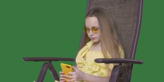 年轻女孩穿着黄色的衣服坐在沙滩椅上，看着手机。长发飘飘的女孩在椅子上放松，绿色背景是智能手机。Alpha通道键控绿色屏幕