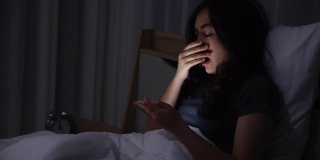 一名亚洲年轻女子晚上躺在床上玩智能手机，在卧室熄灯。在手机上阅读社交。在昏暗的光线下使用手机对眼睛有健康影响。失眠的概念