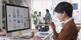 年轻的亚洲开发者使用ai(人工智能)语音识别在移动应用订单智能办公系统，与创意团队在现代办公电脑上工作。Ux UI设计机构