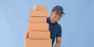 亚洲快乐快递员戴着一顶帽子和一件蓝色衬衫携带纸包盒子孤立在蓝色背景与微笑的脸。邮政投递服务的概念。
