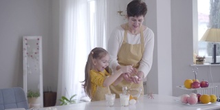 快乐的白人女孩帮助祖母在室内做饭。笑着把鸡蛋打到碗里的小孙女。家庭为忏悔日烤薄饼。生活方式、饮食、忏悔节。