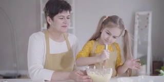 漂亮的小女孩和奶奶坐在厨房里，用面粉打鸡蛋。可爱的白人小孩在忏悔日帮忙做美味的煎饼。烘烤,生活方式,忏悔节。