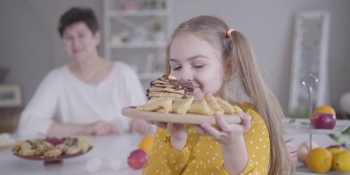 一个快乐的白人小女孩的肖像，闻着烤薄饼，把装有甜点的盘子送给微笑的祖母。焦点从孙女变成了背景中快乐的女人。忏悔节。