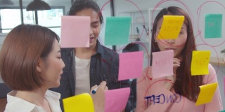 年轻的亚洲女性和她的同事在现代办公室的玻璃墙上用便利贴写想法