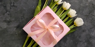 白色郁金香花和粉红色礼盒在深灰色的背景平lay。4K视频俯视图。三月八日母亲节快乐