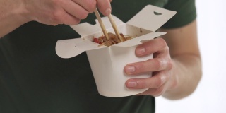 一个年轻人用筷子从白色的纸盒里夹着蔬菜吃中国面条。孤立，在白色背景上