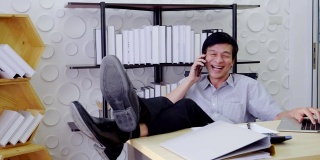 一位亚洲资深人士正坐在办公室里。用手机打电话，说话，工作，笑笑脸，快乐，成功快乐