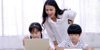 在家教育技术，亚洲年轻的母亲在教家庭作业的儿子和女儿在客厅，小女孩坐在看平板电脑寻找学习信息。