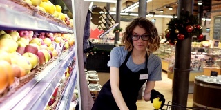 侧面的漂亮的白人工人在黑色围裙和手套stocking在超市的水果。年轻员工在工作。卷曲的女性排列水果在架子上