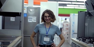 超市商店正面女售货员或售货员肖像。一个穿着蓝色衬衫，带着空吊牌的女人微笑着看着镜头。背景是家用电器
